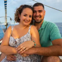 Pareja joven abrazada y sonriente en la proa de un velero, con el mar y el cielo al fondo