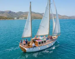 Vista aérea de un velero clásico navegando a vela con pasajeros sobre agua turquesa junto a una costa montañosa