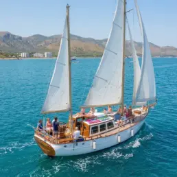Vista aérea de un velero clásico navegando a vela con pasajeros sobre agua turquesa junto a una costa montañosa