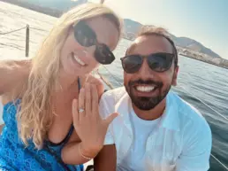 Couple in sunglasses taking a selfie as she shows an engagement ring, sea in the background