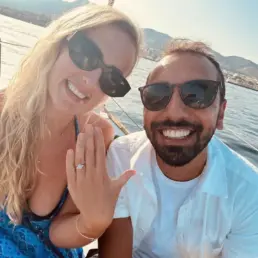 Pareja con gafas de sol haciéndose un selfie mientras ella muestra un anillo de compromiso, mar al fondo