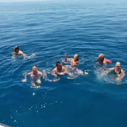 Group of swimmers in the blue sea waving beside the hull of an anchored sailing yacht