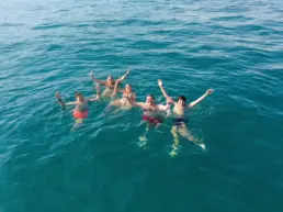 Five people in turquoise water with arms raised celebrating beside the sailing yacht