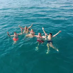 Cinco personas en el agua turquesa con los brazos en alto celebrando junto al velero