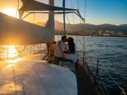 Couple kissing on the bow of a yacht, backlit as the sun sets over the coast