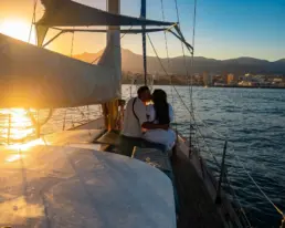Pareja besándose en la proa de un velero a contraluz con el sol poniéndose sobre la costa
