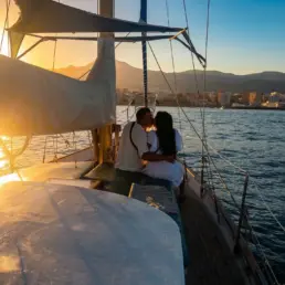 Pareja besándose en la proa de un velero a contraluz con el sol poniéndose sobre la costa