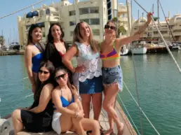 Group of women in swimwear posing on a yacht's deck with Puerto Marina Benalmádena behind