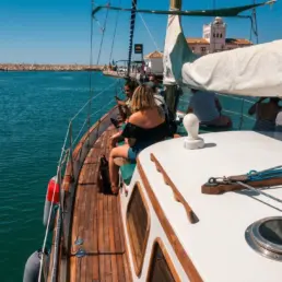 Pasajeros sentados a lo largo de la borda de teca de un velero mirando el mar al salir del puerto