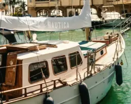 Detalle de un velero clásico amarrado con casco blanco, línea de teca y defensas sobre el agua del puerto