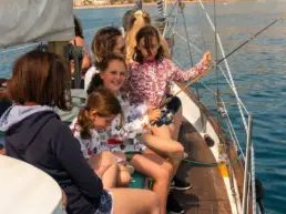 People seated on a yacht's rail, one holding a fishing rod, with the coast behind