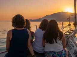 Four women seen from behind at a yacht's stern watching the golden sunset over the mountains