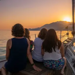 Four women seen from behind at a yacht's stern watching the golden sunset over the mountains