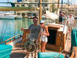 Couple posing with thumbs up at a yacht's helm with Puerto Marina Benalmádena behind