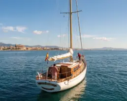 Vista aérea apaisada de un velero con la vela recogida y bandera española, ciudad costera al fondo