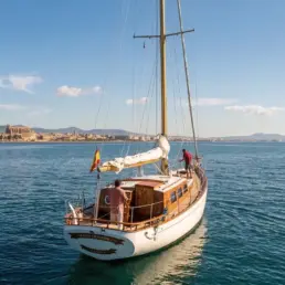 Vista aérea apaisada de un velero con la vela recogida y bandera española, ciudad costera al fondo