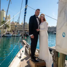 Pareja de novios vestidos de boda posando en la proa de un velero con Puerto Marina de Benalmádena al fondo