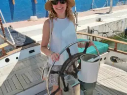 Woman in a hat and white dress posing at the helm of a yacht in the harbour