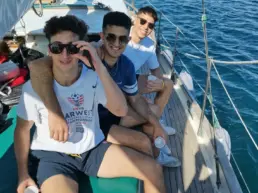 Three young men in sunglasses with drinks seated on a yacht's deck as it leaves the harbour