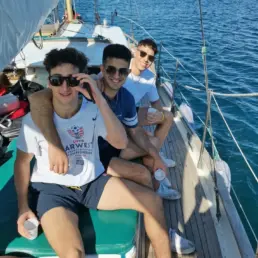 Three young men in sunglasses with drinks seated on a yacht's deck as it leaves the harbour
