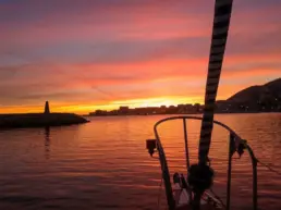 Orange and mauve sunset over a lighthouse and the lit coastal skyline, rigging in silhouette