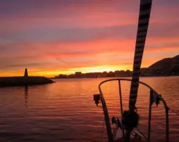 Orange and mauve sunset over a lighthouse and the lit coastal skyline, rigging in silhouette