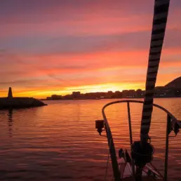Atardecer de cielo naranja y malva sobre un faro y el skyline iluminado de la costa, aparejo en silueta