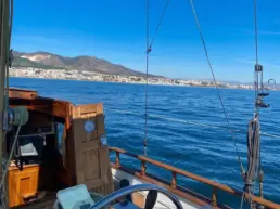 Skipper's hands steering a yacht's teak wheel with the coast and blue sky behind