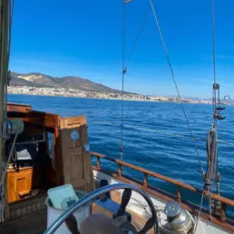 Manos del patrón gobernando el timón de teca de un velero con la costa y el cielo azul al fondo