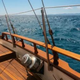 Detalle de un winch cromado y aparejo sobre la borda de teca de un velero con el mar abierto al fondo
