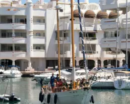 Yacht manoeuvring in the Puerto Marina Benalmádena basin with white buildings and a group aboard