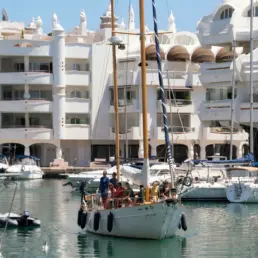 Yacht manoeuvring in the Puerto Marina Benalmádena basin with white buildings and a group aboard