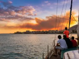Three people seen from behind on a yacht's bow watching a sunset of orange clouds