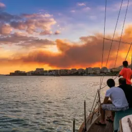 Tres personas de espaldas en la proa de un velero contemplando un atardecer de nubes naranjas