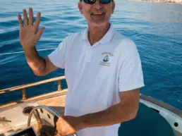 Skipper in sunglasses and white polo waving at a yacht's helm with the coast behind