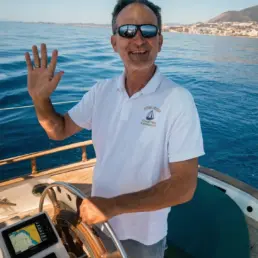 Patrón con gafas de sol y polo blanco saludando al timón de un velero con la costa al fondo