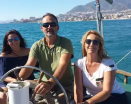Patrón al timón de un velero entre dos mujeres con gafas de sol y el mar turquesa al fondo