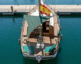 Top-down view of a moored yacht with a teak deck, green cushions, wheel and Spanish flag