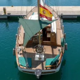 Vista cenital de un velero amarrado con cubierta de teca, cojines verdes, timón y bandera española