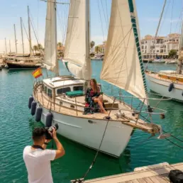 Fotógrafo en el pantalán retratando a una modelo en un velero con las velas izadas en el puerto