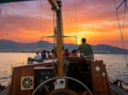 Group on a yacht's deck watching a red sunset behind the mountains, wheel in the foreground