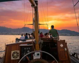 Grupo en la cubierta de un velero contemplando un atardecer rojo tras las montañas, timón en primer plano