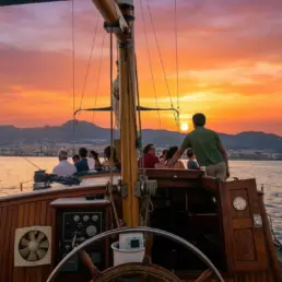 Grupo en la cubierta de un velero contemplando un atardecer rojo tras las montañas, timón en primer plano