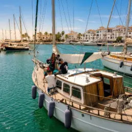 Pasajeros a bordo de un velero amarrado en un puerto con palmeras y edificios blancos