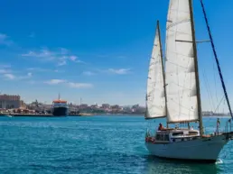 Yacht sailing with white sails raised over turquoise water off a coastal city with a cathedral