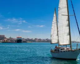 Velero navegando con las velas blancas izadas sobre agua turquesa frente a una ciudad costera