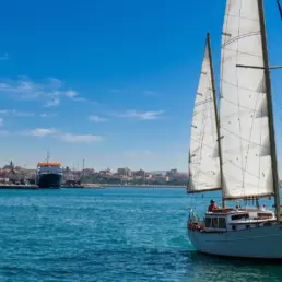 Velero navegando con las velas blancas izadas sobre agua turquesa frente a una ciudad costera