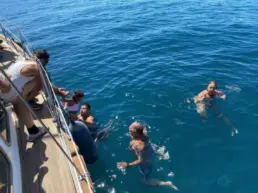 Swimmers beside a yacht's hull while others watch from the teak rail