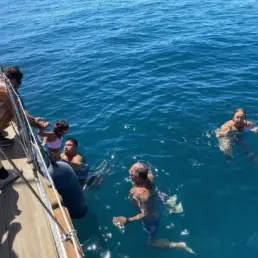 Bañistas nadando junto al casco de un velero mientras otros observan desde la borda de teca