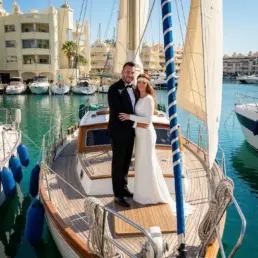 Pareja de novios de pie en la proa de un velero con los edificios de Puerto Marina de Benalmádena al fondo